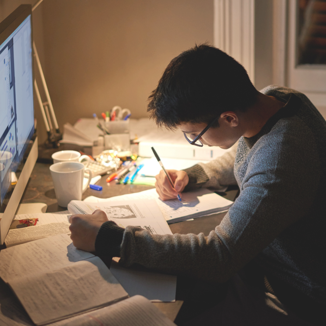 A person wearing glasses sits at a desk in a warmly lit room, writing in a notebook with a pen. An open computer monitor, books, and notebooks are visible on the desk.